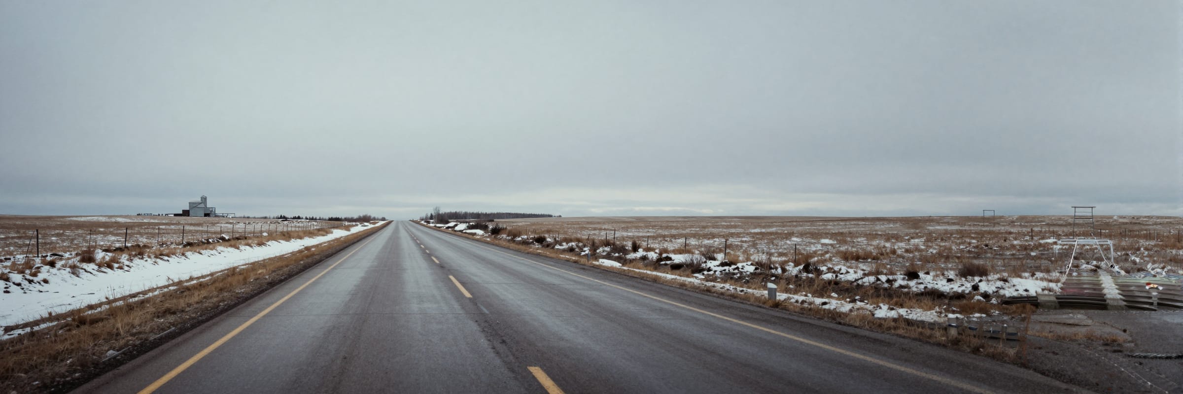 A two-lane Alberta highway running straight to a flat horizon between snow-crusted fields.