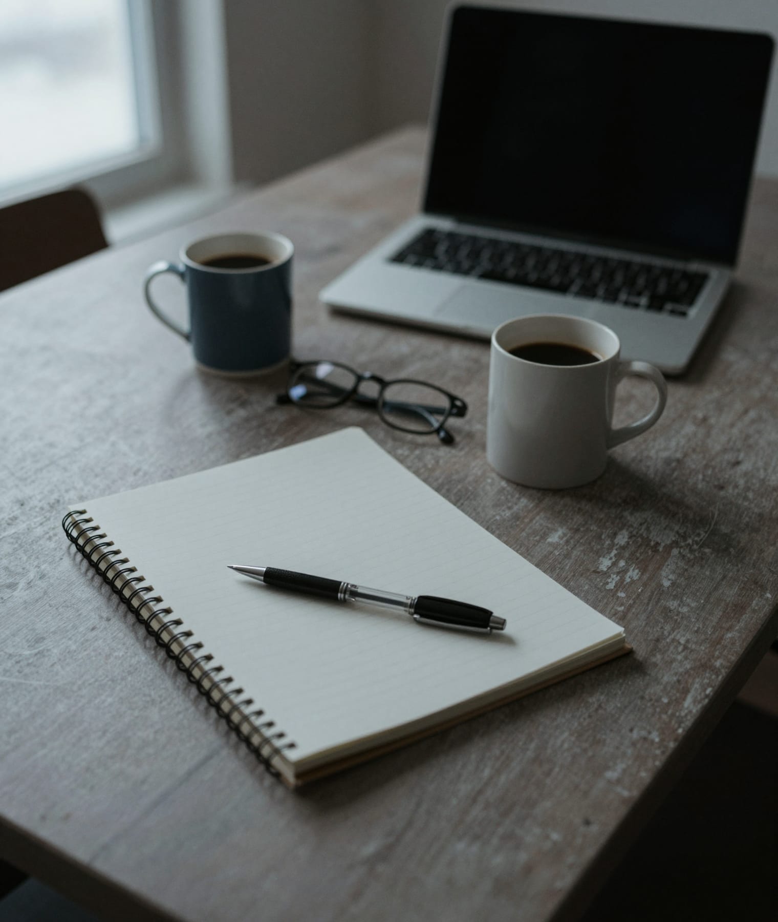 A meeting table with an open notebook, a pen, two mugs of coffee and a closed laptop.