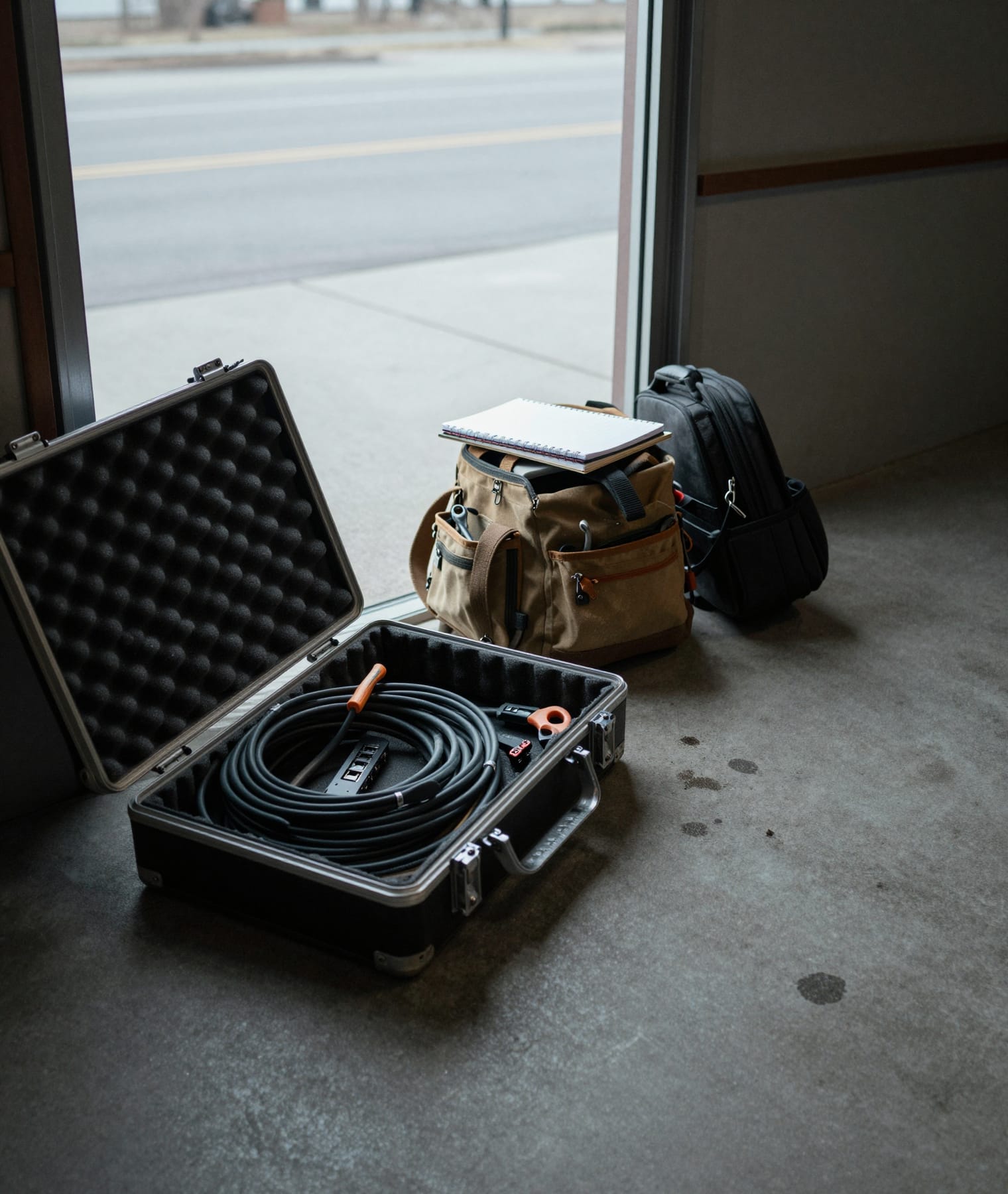 An equipment case open on a concrete floor beside a toolbag and a laptop bag, just inside an open door.