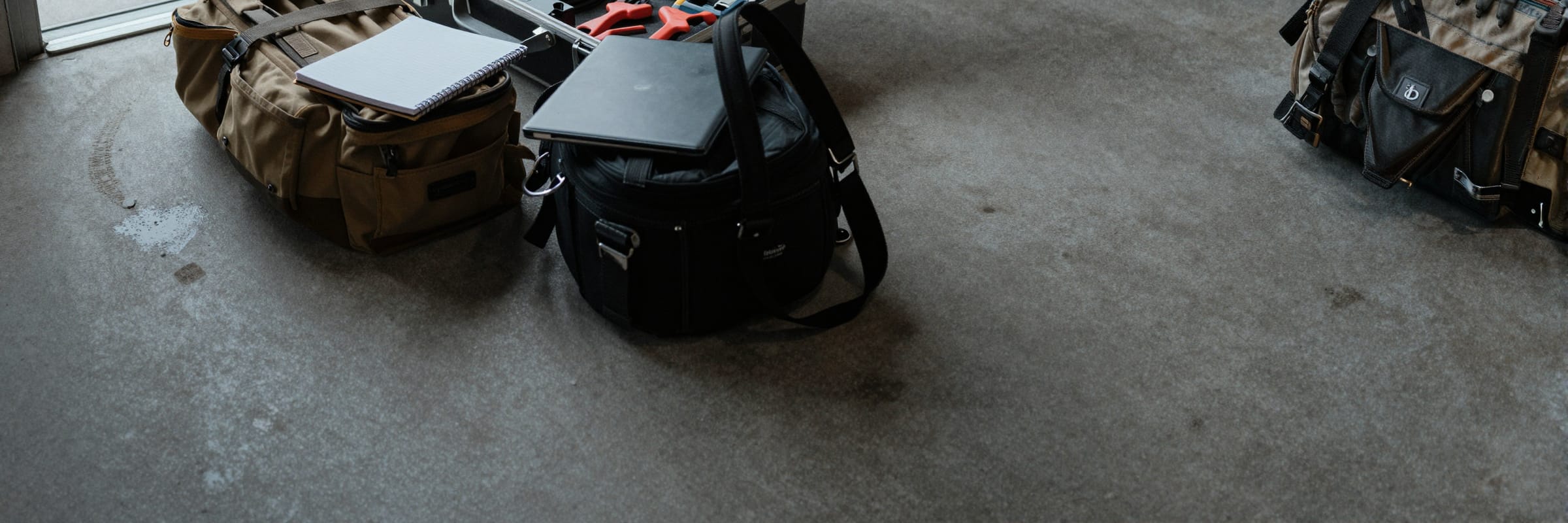 An equipment case open on a concrete floor beside a toolbag and a laptop bag, just inside a roll-up door.