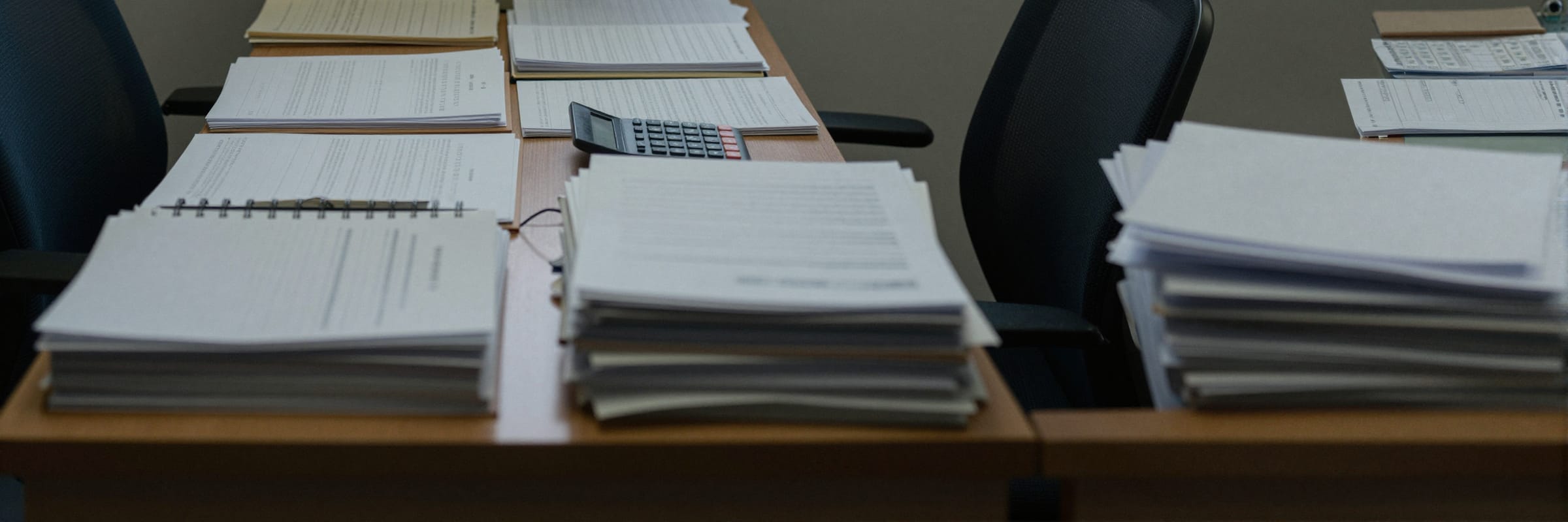 A desk covered edge to edge in stacked paper and open documents under a grey window.